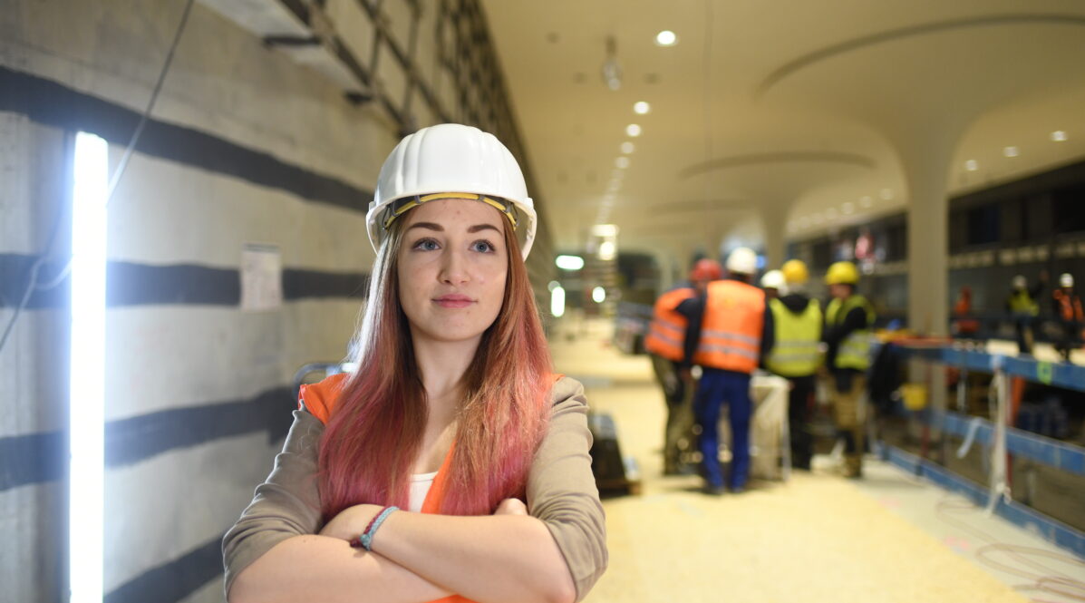 Ingenieurin Svenja steht mit verschränkten Armen auf einer Baustelle. Hinter ihr sind ihre Kolleg*innen in Schutzkleidung zu sehen.