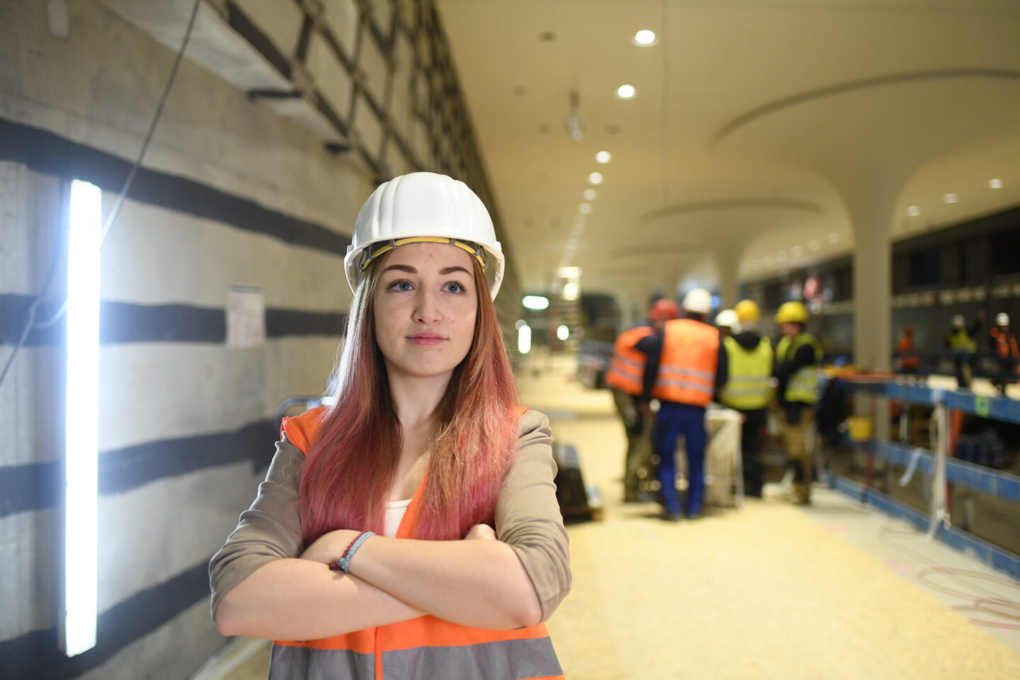 Ingenieurin Svenja steht mit verschränkten Armen auf einer Baustelle. Hinter ihr sind ihre Kolleg*innen in Schutzkleidung zu sehen.