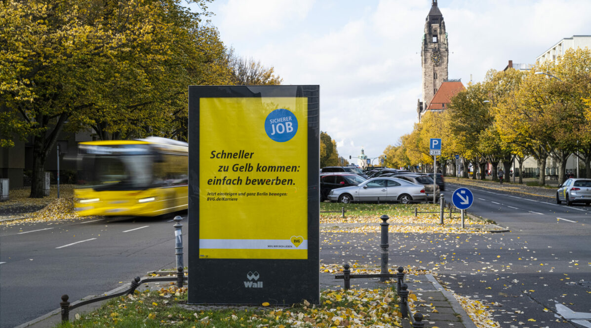 In der Mittelinsel einer Straße steht eine Außenwerbung für BVG-Karriere. Im Hintergrund steht ein gelber BVG-Bus.