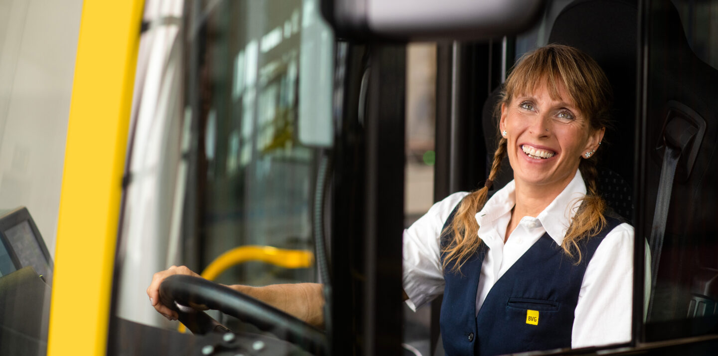 Busfahrerin Manuela sitzt am Steuer eines BVG-Busses und schaut lächelnd durch die Glasscheibe.