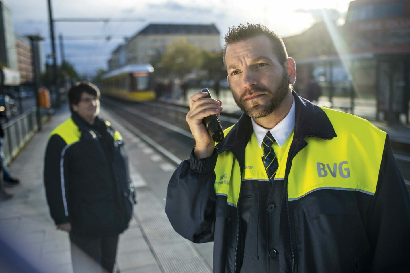 Eine Kollegin und ein Kollege aus dem Bereich Sicherheitsdienst stehen in Schutzkleidung mit Funkgerät am Bahnsteig der Straßenbahn.