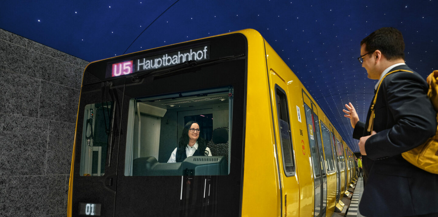 Eine BVG-U-Bahn fährt auf der Station Museumsinsel ein. Die Bahn wird von einer Fahrerin gelenkt. hr Kollege steht auf dem Bahnsteig und winkt ihr zu.