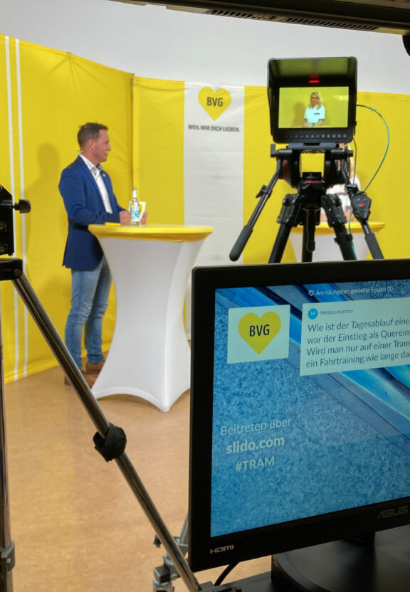 Zu sehen ist das Sendestudio eines Recruiting-Events. Im Vordergrund ein Flachbildschirm und im Hintergrund steht der Moderator vor einer gelben Wand.