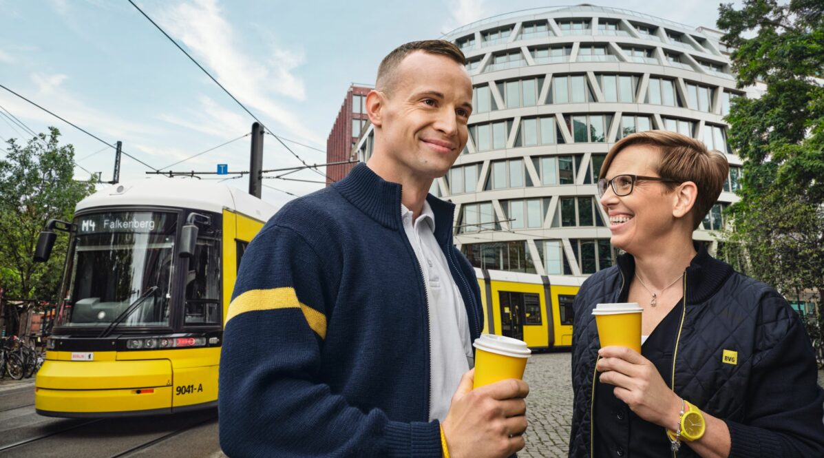 Zwei BVG Mitarbeiter*innen in Fahrdienstkleidung unterhalten sich am Straßenrand. Sie halten gelbe Pappbecher in der Hand. Im Hintergrund fährt die Straßenbahn M4 Falkenberg vorbei.
