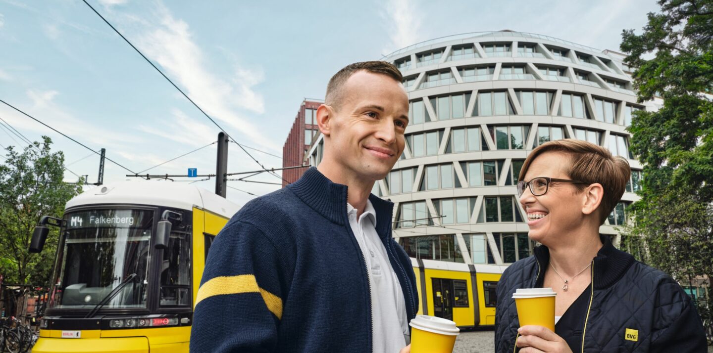 Zwei BVG Mitarbeiter*innen in Fahrdienstkleidung unterhalten sich am Straßenrand. Sie halten gelbe Pappbecher in der Hand. Im Hintergrund fährt die Straßenbahn M4 Falkenberg vorbei.