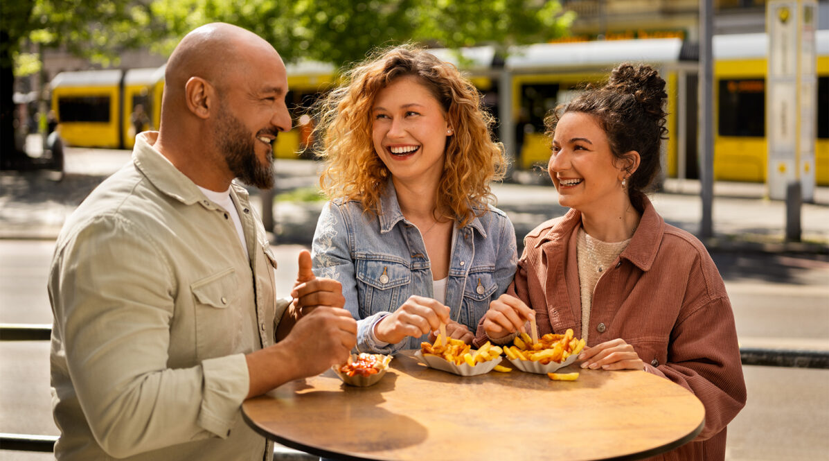3 junge Personen stehen an einem Stehtisch. Sie essen Pommes mit Ketchup. Sie lachen sich zu. Im Hintergrund ist eine Straßenbahn zu sehen. Und eine Haltestelle der BVG.