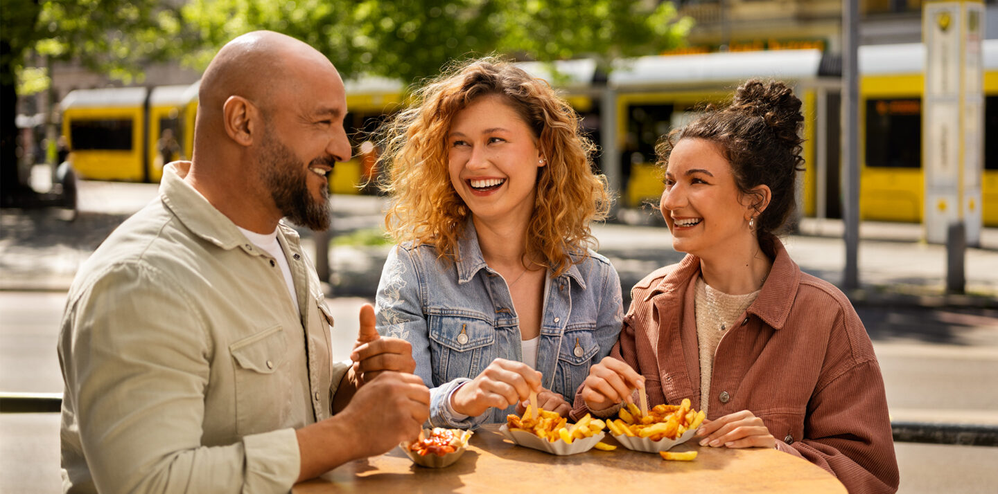 3 junge Personen stehen an einem Stehtisch. Sie essen Pommes mit Ketchup. Sie lachen sich zu. Im Hintergrund ist eine Straßenbahn zu sehen. Und eine Haltestelle der BVG.