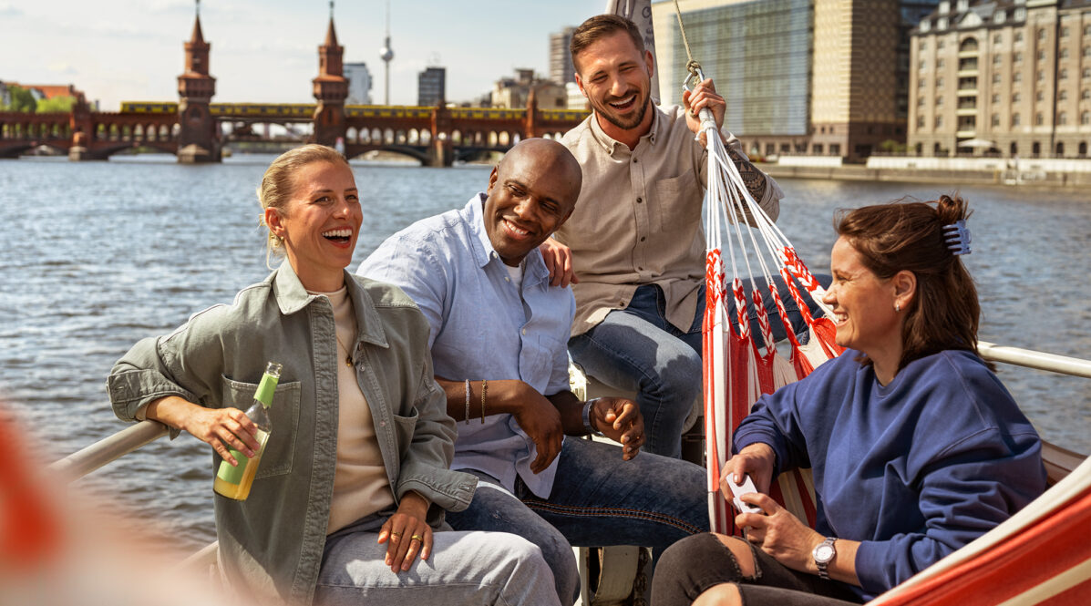 Personen sitzen in Freizeitkleidung in einem Boot. Sie lachen. Im Hintergrund sind Wasser, eine Brücke und Wahrzeichen von Berlin zu sehen.
