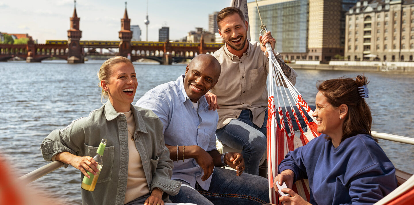Personen sitzen in Freizeitkleidung in einem Boot. Sie lachen. Im Hintergrund sind Wasser, eine Brücke und Wahrzeichen von Berlin zu sehen.