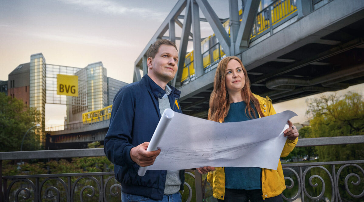 Ein Mann und eine Frau halten einen Bauplan in der Hand. Sie stehen auf einer Brücke. Im Hintergrund ist ein U-Bahn-Viadukt der BVG mit einem großen rechteckigen BVG-Logo zu sehen. Eine U-Bahn fährt auf dem Viadukt.