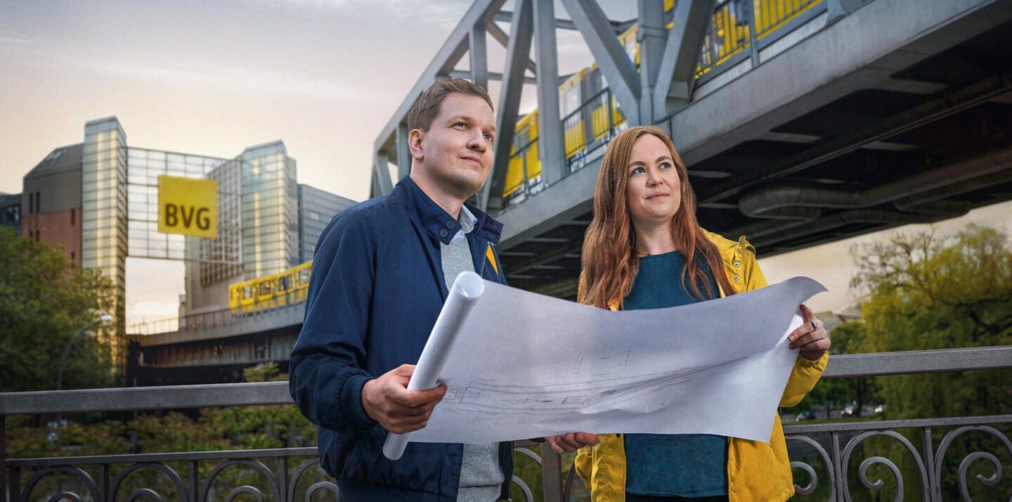 Ein Mann und eine Frau halten einen Bauplan in der Hand. Sie stehen auf einer Brücke. Im Hintergrund ist ein U-Bahn-Viadukt der BVG mit einem großen rechteckigen BVG-Logo zu sehen. Eine U-Bahn fährt auf dem Viadukt.