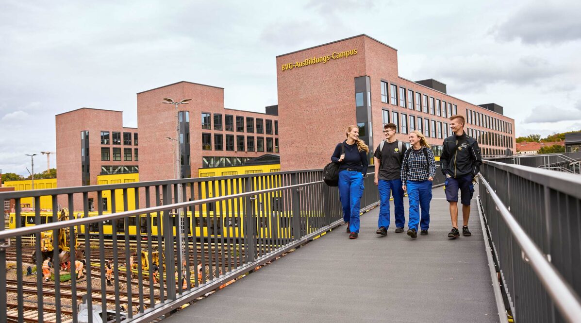 Vier Azubis laufen über eine Brücke, im Hintergrund sieht man das neue Ausbildungszentrum