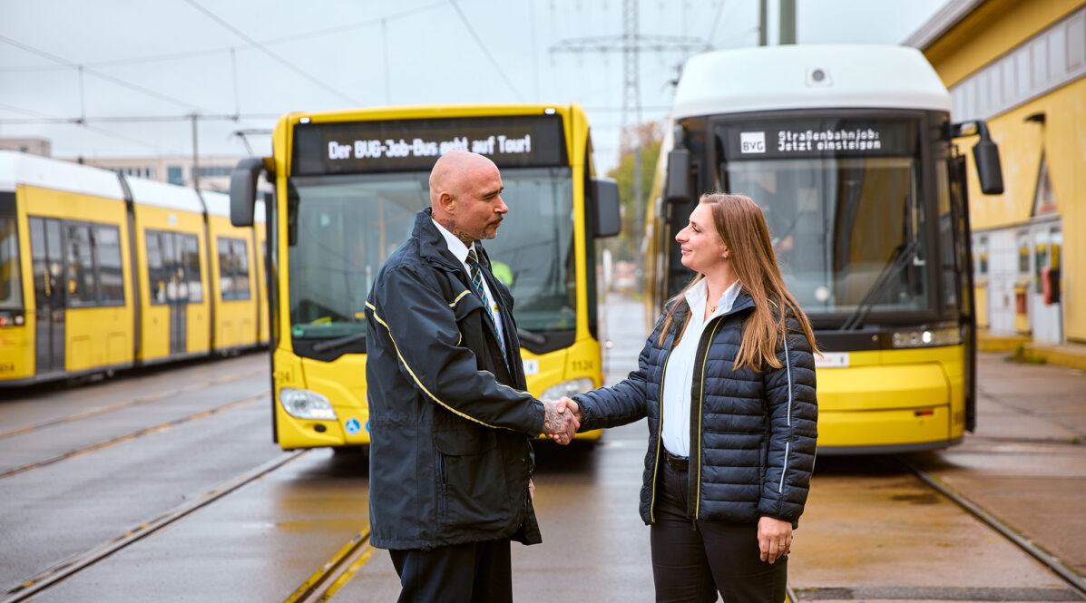 Busfahrer und Tramfahrerin geben sich die Hand. Im Hintergrund sieht man einen Bus und eine Straßenbahn.