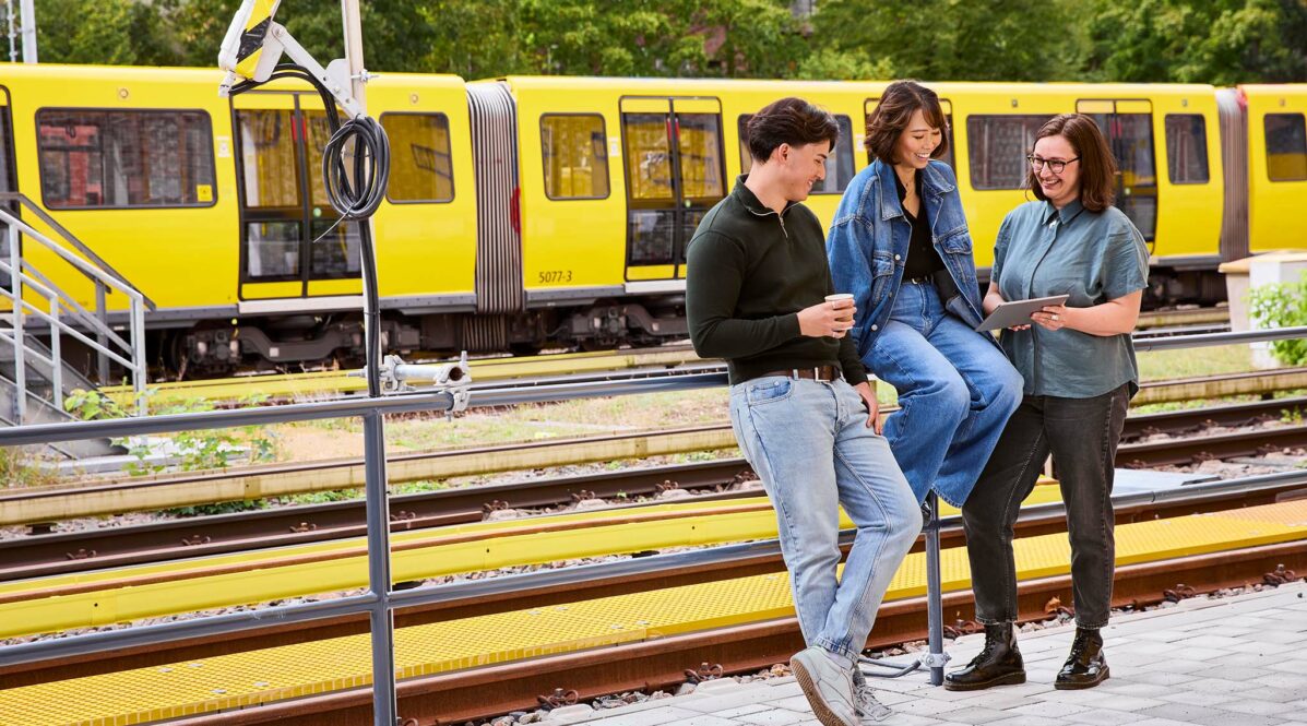 Drei Personen sitzen auf einem Geländer und machen Pause. Im Hintergrund sieht man eine U-Bahn.