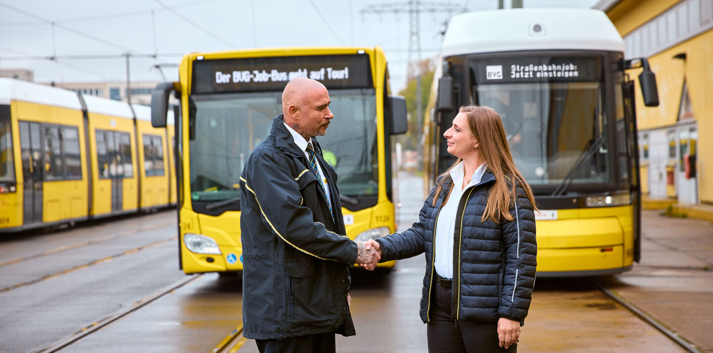 Ein Mann und eine Frau stehen vor einem Bus und einer Straßenbahn und geben sich die Hand