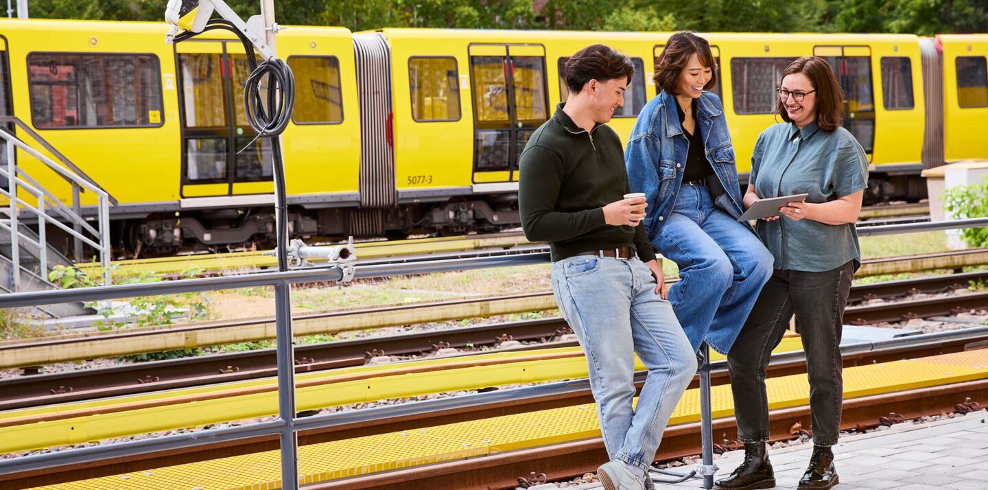 Drei Personen sitzen auf einem Geländer und machen Pause. Im Hintergrund sieht man eine U-Bahn.