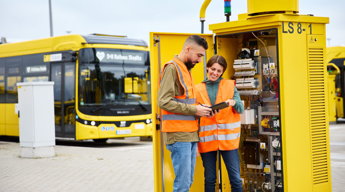 Ein Ingenieur und eine Ingenieurin stehen vor einer Ladesäule. Im Hintergrund steht ein E-Bus