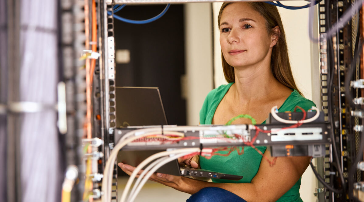 Frau sitzt mit Laptop im Serverraum, Kabel sind zu sehen.